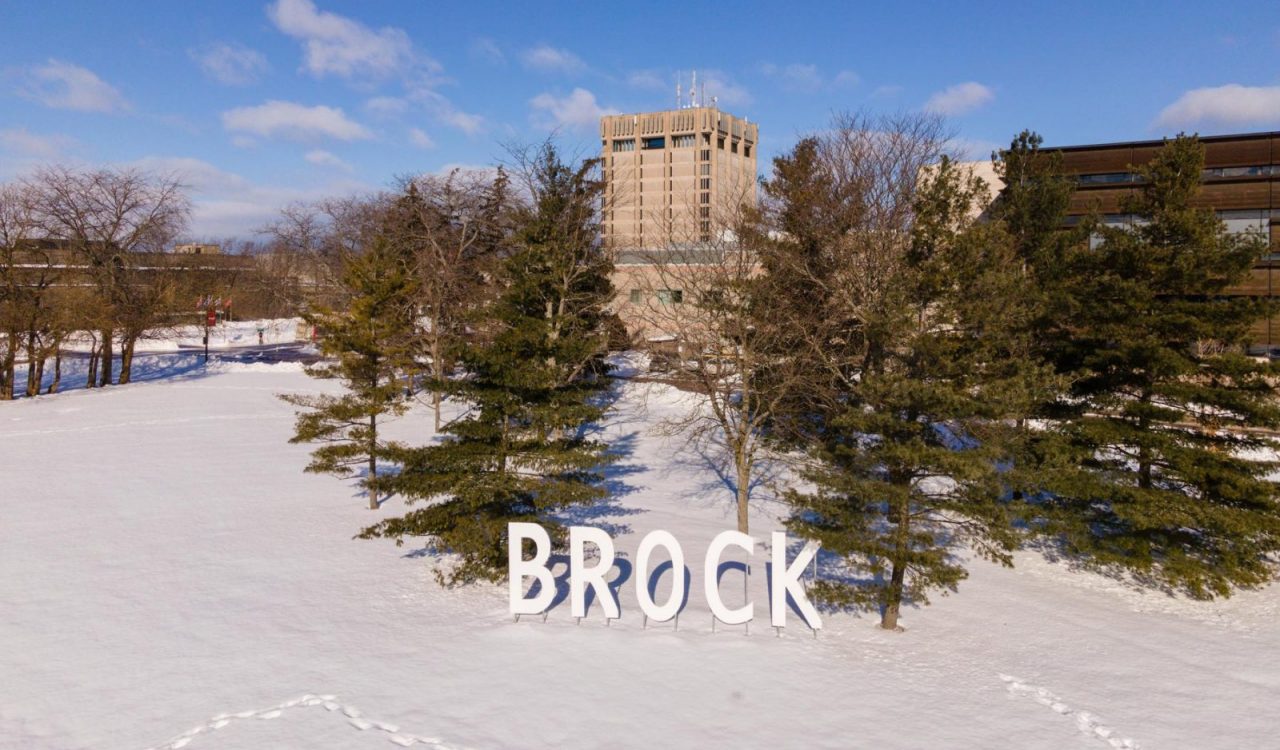 Winter scene of the Brock tower in the background and in the foreground, pine trees and giant lettering forming the word “Brock”