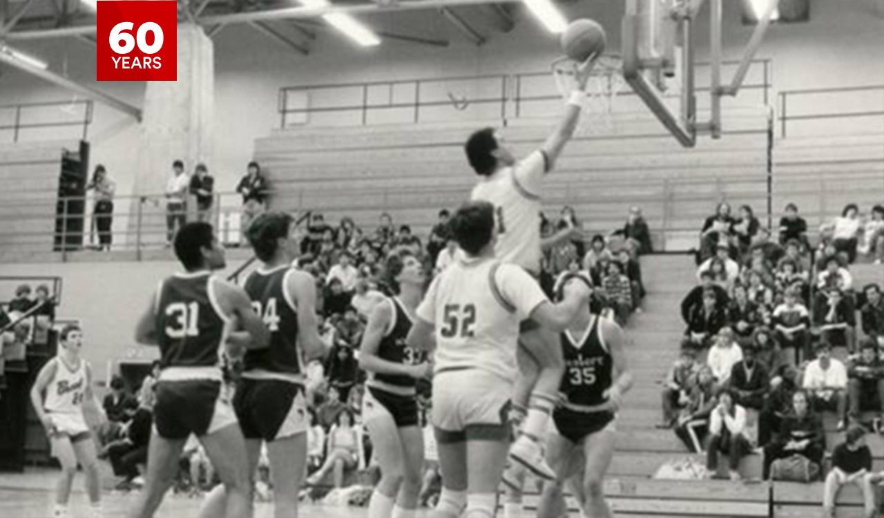 A black-and-white photograph of a basketball game in progress inside a gymnasium. A player in a light-colored uniform is mid-air, attempting a layup, while players from both teams in light and dark uniforms are gathered around, watching or preparing to rebound. Spectators fill the bleachers in the background, attentively watching the action.