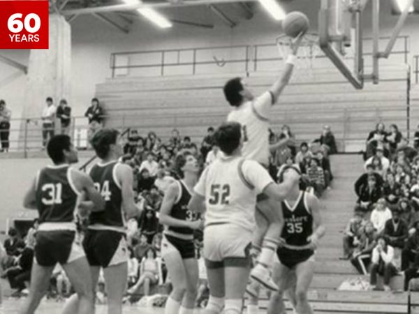 A black-and-white photograph of a basketball game in progress inside a gymnasium. A player in a light-colored uniform is mid-air, attempting a layup, while players from both teams in light and dark uniforms are gathered around, watching or preparing to rebound. Spectators fill the bleachers in the background, attentively watching the action.