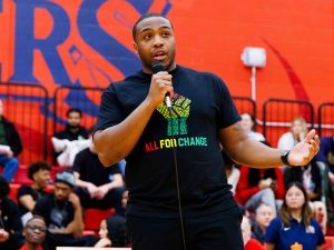 A speaker addresses the crowd at a Brock University event, wearing a black "All For Change" T-shirt featuring a raised fist design in red, yellow, and green. The audience in the background listens attentively in the gymnasium, surrounded by Brock Badgers branding.