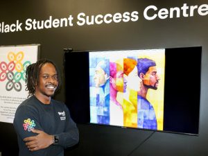 Justin Mafie stands in front of a video screen displaying a brightly coloured image of four Black individuals.