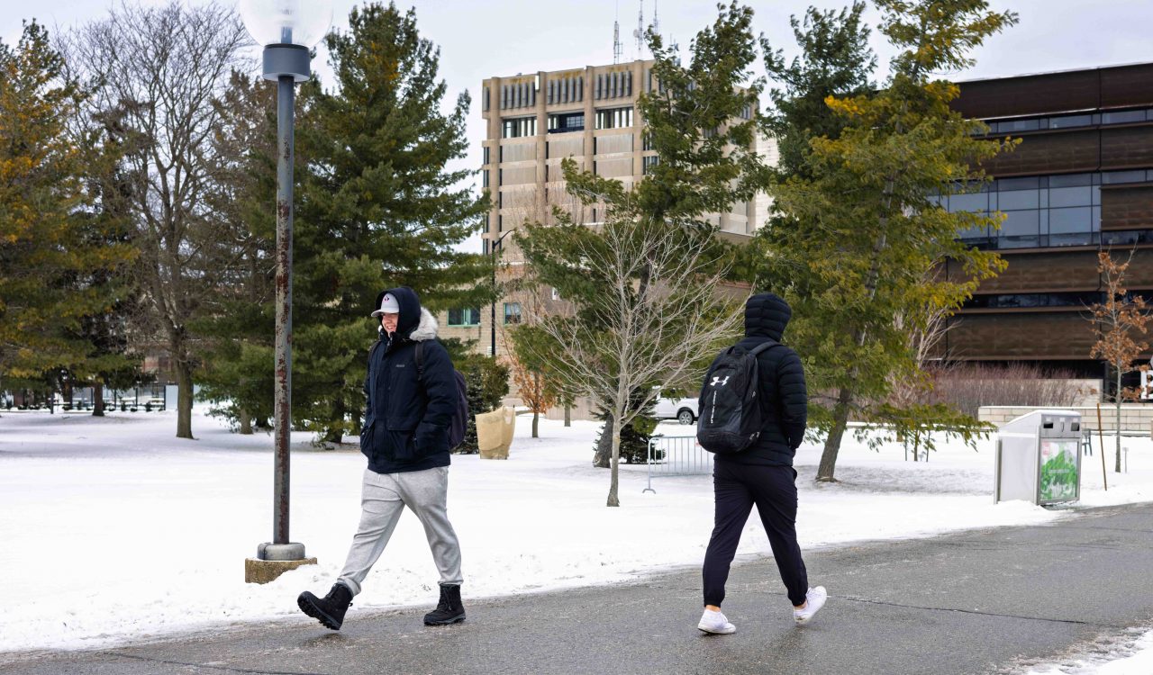 Two people bundled up in heavy coats pass each other on an outdoor pathway during winter at Brock University’s main campus. There are buildings and green trees in the background.