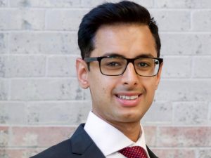 Headshot of Arjun Rijhwani wears a suit and standing in front of a grey and red brick wall.