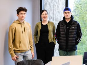 Three people pose for a photo in front of a window in a small university classroom.