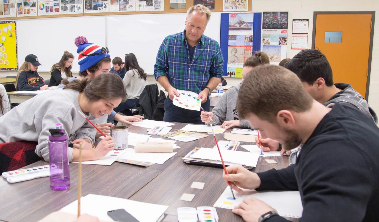 A man stands at a table surrounded by university students painting and making art.