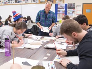 A man stands at a table surrounded by university students painting and making art.