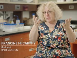 Screen grab of documentary picturing Francine McCarthy sitting on a chair, looking at the camera and gesturing with both arms, with the title “Francine McCarthy, Geologist, Brock University” appearing on screen to her left.