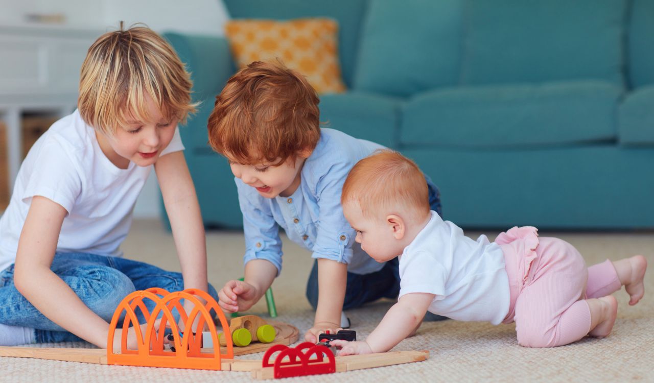 An infant, toddler and young child play together on the floor of a living room.