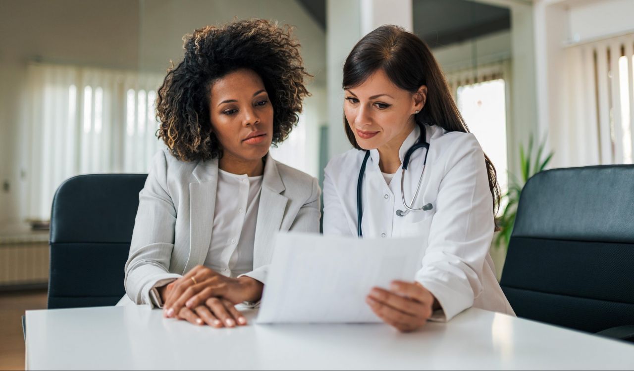 Two women, one in a suit and one in a lab coat with a stethoscope around her neck, sit at a table reviewing a document.