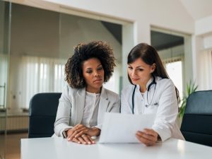 Two women, one in a suit and one in a lab coat with a stethoscope around her neck, sit at a table reviewing a document.