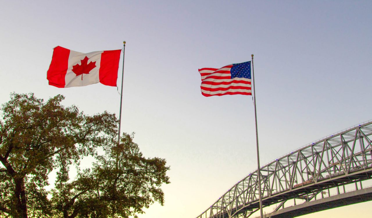 The Canadian and American flags fly against a sunset sky with a bridge in the background.