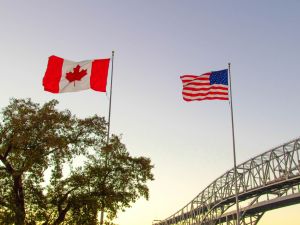 The Canadian and American flags fly against a sunset sky with a bridge in the background.