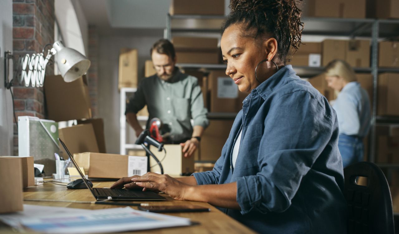 Woman in the foreground sitting at a counter typing on a laptop with parcels and papers around her, with a man and a woman examining boxes and a shelf of boxes fuzzed out in the background.