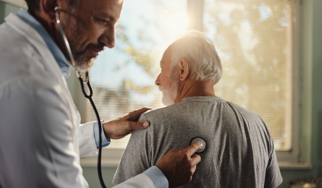 A doctor holds a stethoscope up to the back of an elderly man.