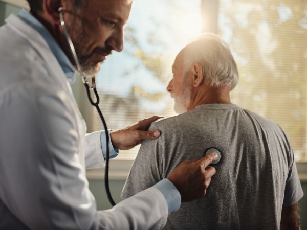 A doctor holds a stethoscope up to the back of an elderly man.