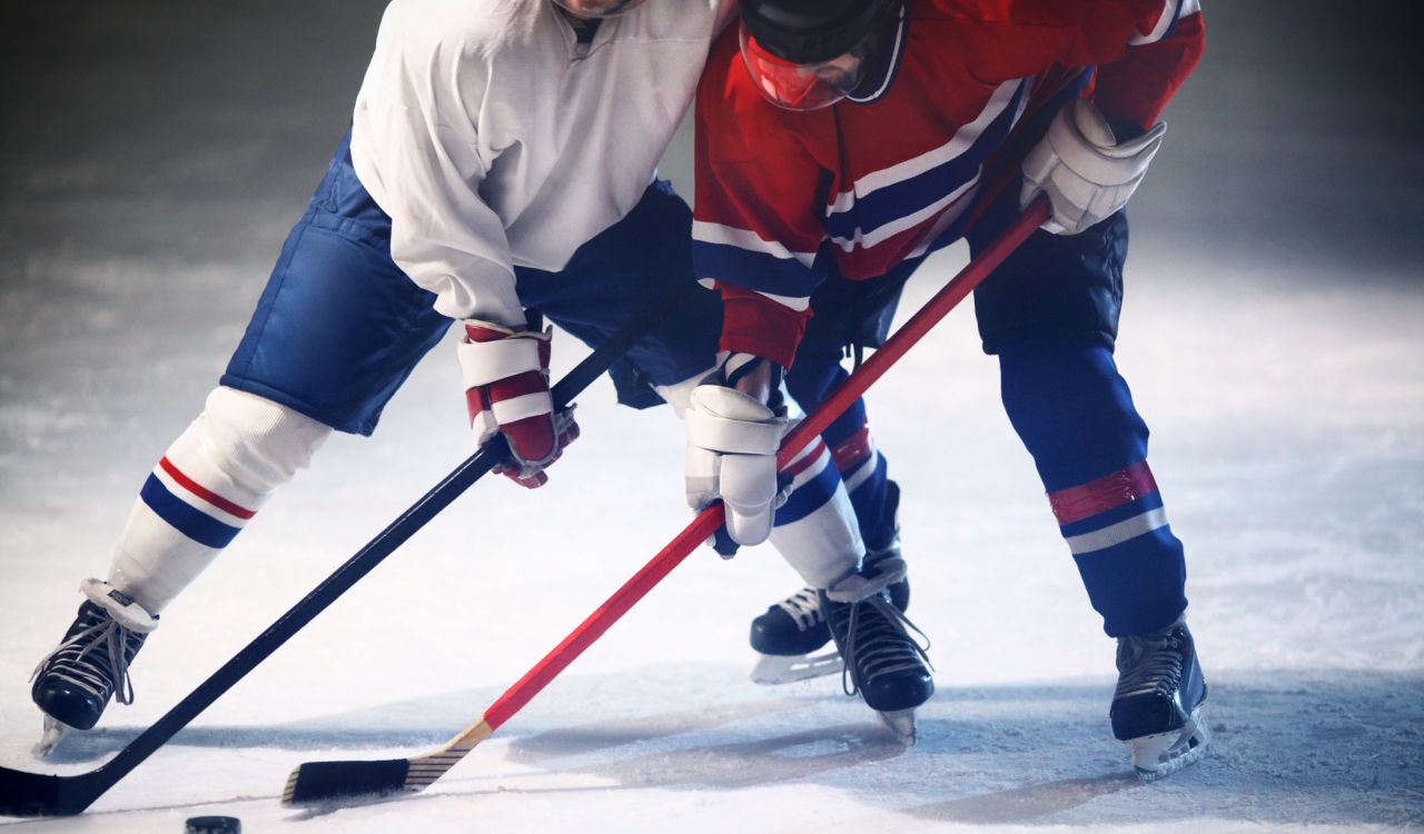 Two unrecognizable adult men reaching for the puck with their hockey sticks while playing ice hockey.