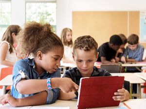 Two elementary school children work together on a tablet while a teacher helps other children in the background.