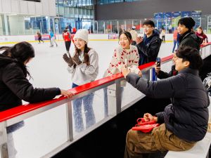 A group of friends enjoys a day at the ice rink, smiling, clapping and capturing moments on their phones while others skate in the background. Some are on the ice in winter attire and others sit on benches along the rink, chatting and relaxing.