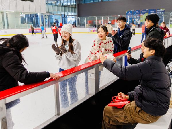 A group of friends enjoys a day at the ice rink, smiling, clapping and capturing moments on their phones while others skate in the background. Some are on the ice in winter attire and others sit on benches along the rink, chatting and relaxing.