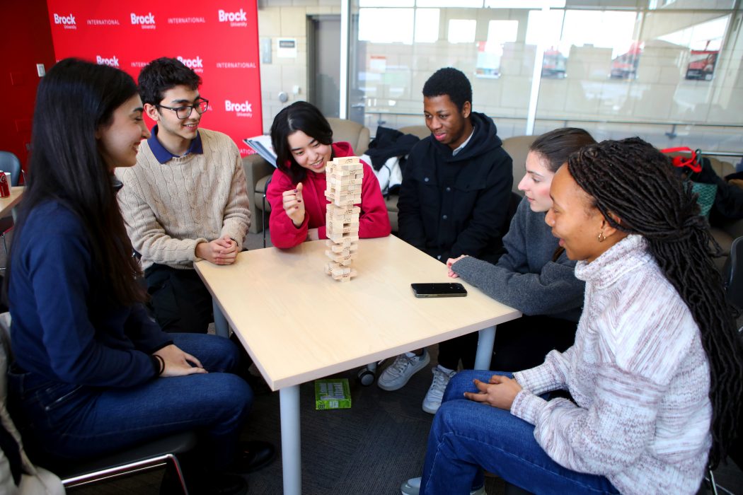 Six university students sit at a table in a meeting room playing a game of jenga on a table.