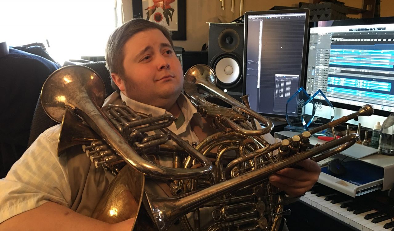 A man sits at a computer desk in front of sound editing software holding a large brass instrument.