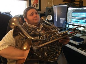 A man sits at a computer desk in front of sound editing software holding a large brass instrument.