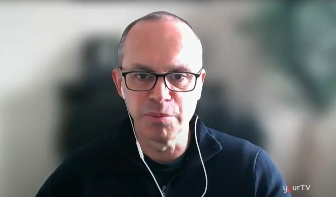 Louis Volante wears a blue sweater and headphones and takes part in a virtual television interview.