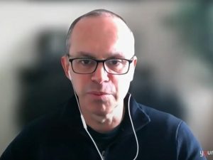 Louis Volante wears a blue sweater and headphones and takes part in a virtual television interview.