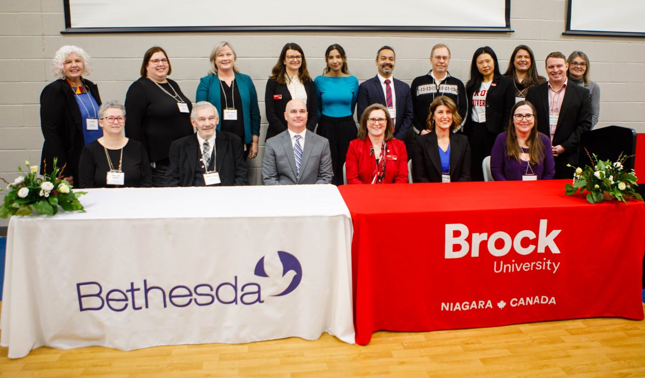 A group of 14 men and women stand posing with some seated at tables with branding which read Bethesda and Brock University in a gymnasium.