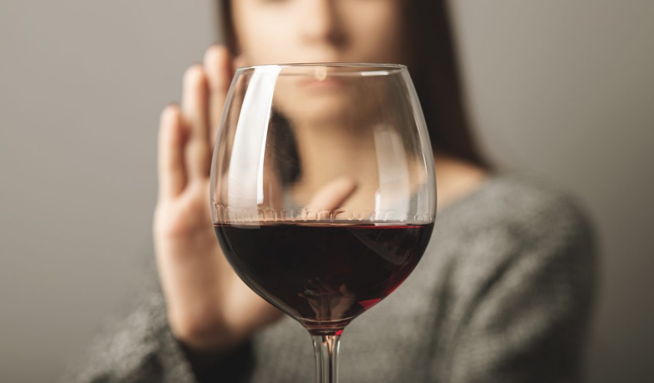 Close-up of a glass of red wine with a woman's hand held up to refuse it in the background.