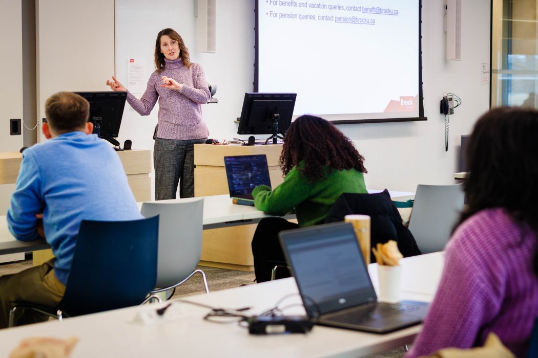 A woman stands speaking to a class of people sitting at desks.