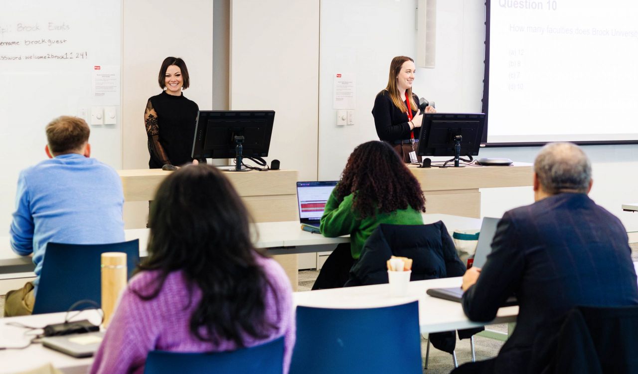 Two women stand at podiums in front of a seated group of people listening attentively in a brightly lit room with white walls and whiteboards.