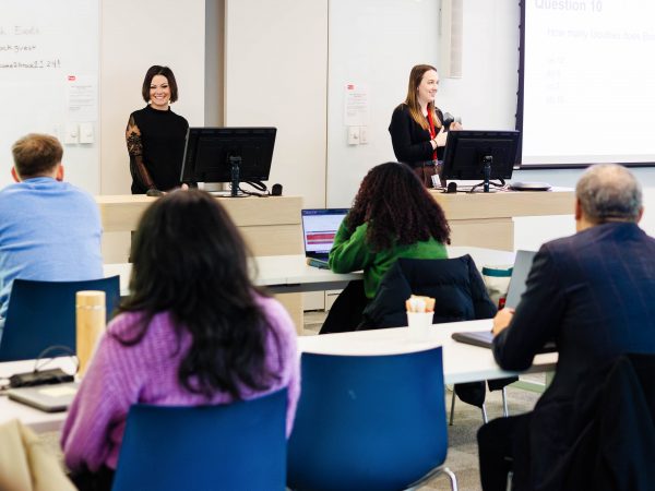 Two women stand at podiums in front of a seated group of people listening attentively in a brightly lit room with white walls and whiteboards.