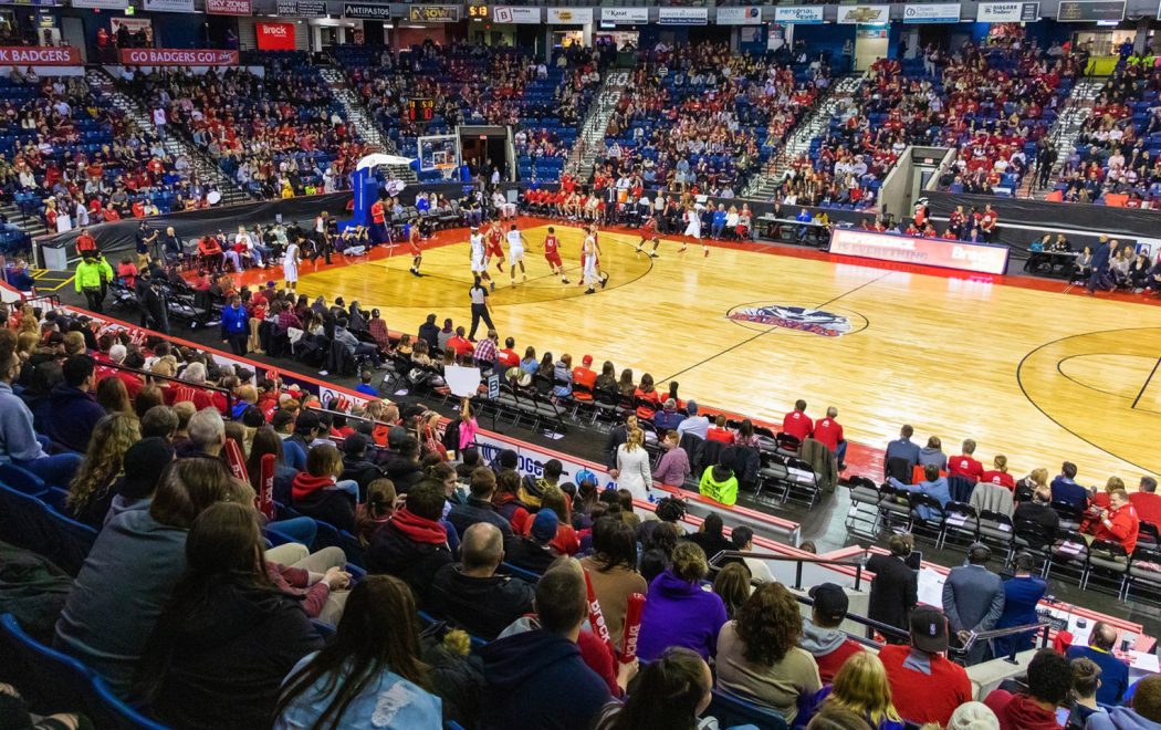 A packed basketball arena shows fans in red cheering as two teams compete on the court. Players are in motion, and spectators fill the stands, creating an electric atmosphere. Coaches, team staff, and officials are visible on the sidelines, with banners and signage supporting the home team displayed prominently.