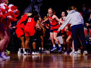 Members of the Brock University women’s basketball team, all wearing red uniforms, smile and cheer as a teammate runs onto the court before a game.