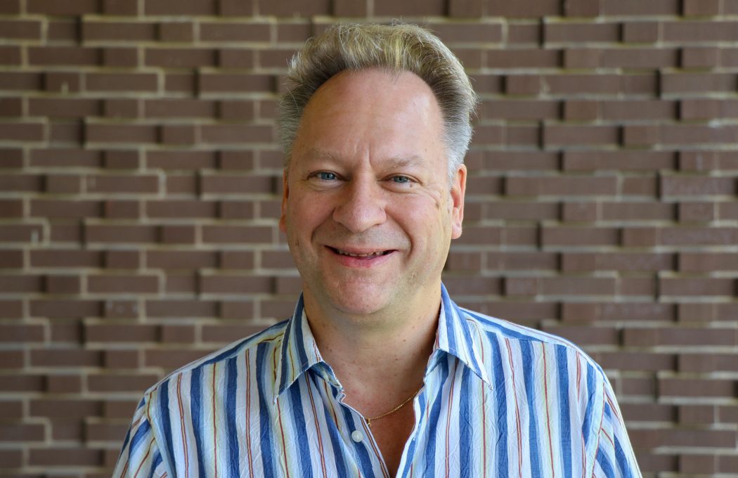 A man smiles warmly at the camera in front of a brick wall background.