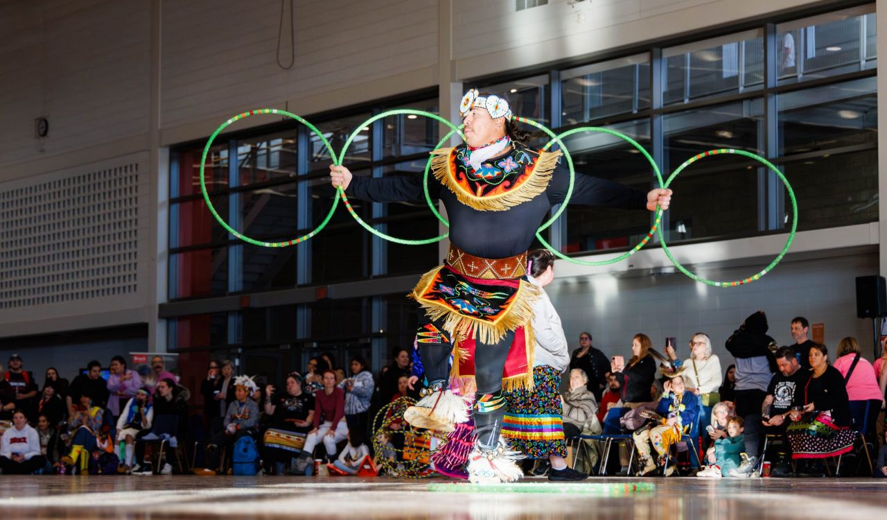 A man in Indigenous regalia performs a hoop dance in front of an audience in a university gymnasium.