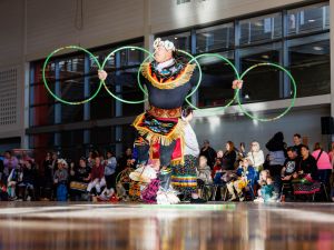 A man in Indigenous regalia performs a hoop dance in front of an audience in a university gymnasium.