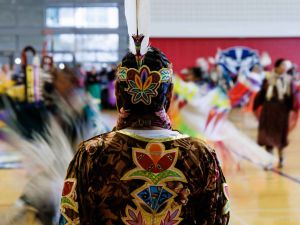 The back of a person wearing Indigenous regalia standing still in the middle of a group of Indigenous dancers who are out of focus because of motion blur.