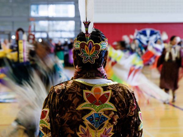 The back of a person wearing Indigenous regalia standing still in the middle of a group of Indigenous dancers who are out of focus because of motion blur.