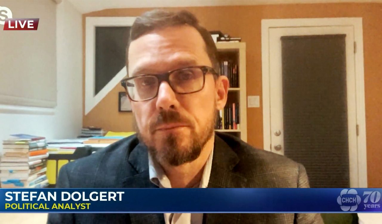 Stefan Dolgert does a digital television interview with books and a door in the background.