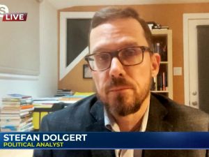 Stefan Dolgert does a digital television interview with books and a door in the background.