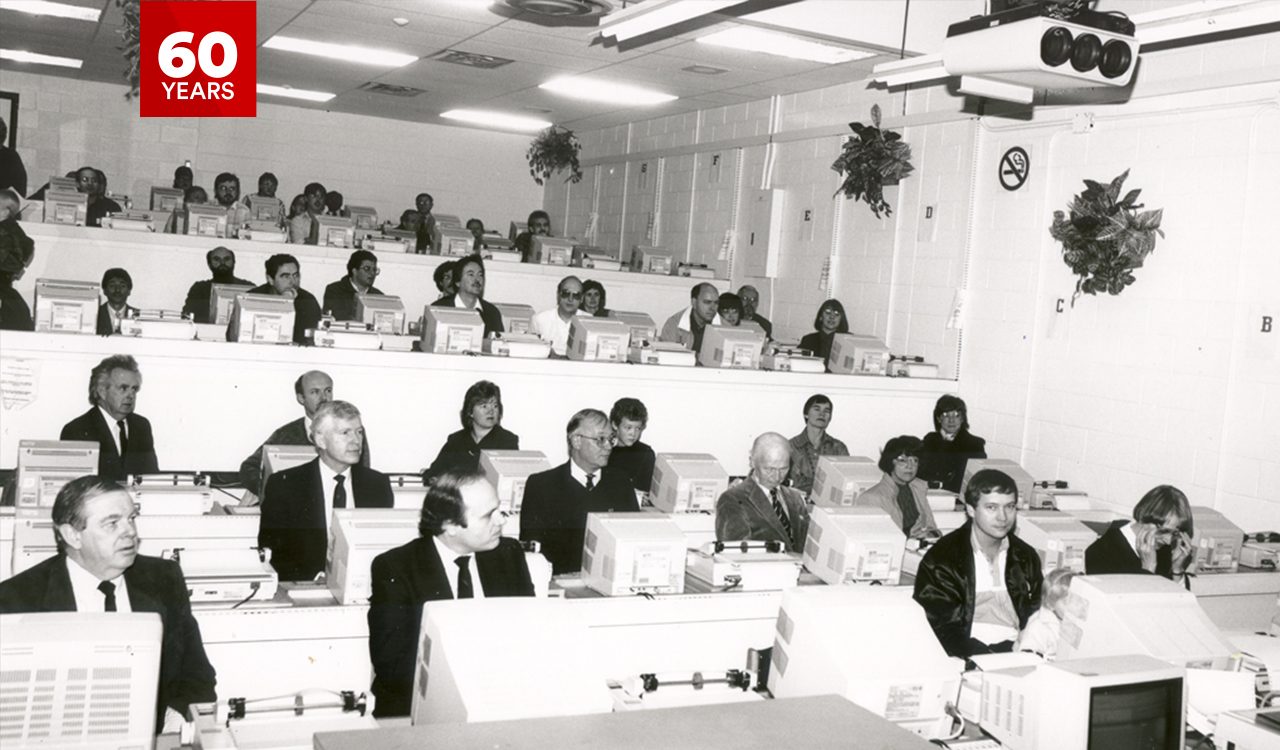 A vintage computer lab or training room from the late 1980s with dated computers and monitors. A professional setting with attendees in formal attire in an academic environment. The room has tiered seating, and the presence of hanging plants and a "no smoking" sign gives it a distinct look.