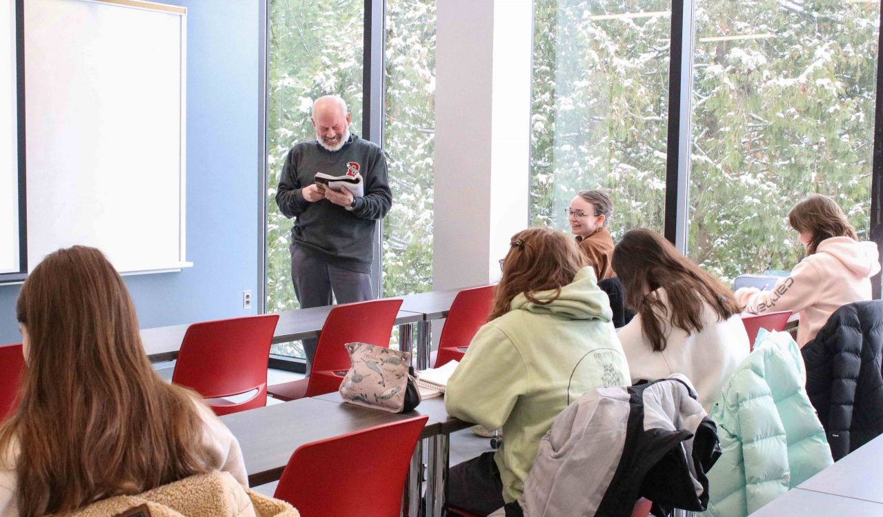 A man stands at the front of a classroom smiling and reading a book in front of a small group of students laughing while looking down at their own text.