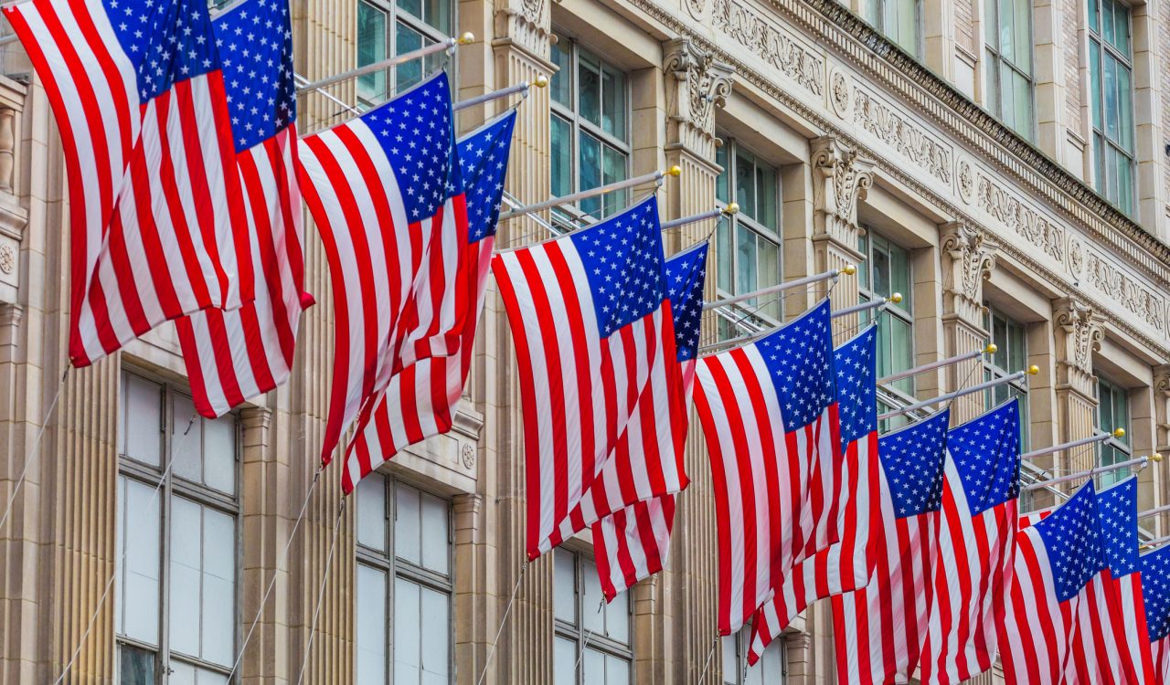 A row of American flags hang from a stone building.