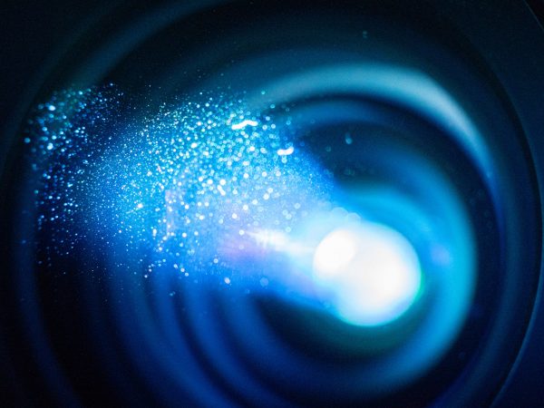Dust sparkles in the bluish light of a 4K projector lens shown in close-up.
