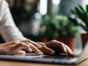 Close-up of hands typing on a laptop that's resting on a table, with green potted plants fuzzed out in the background.