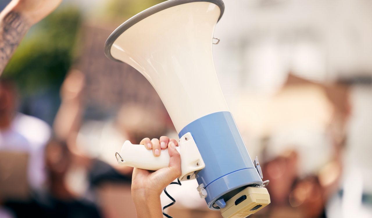 Close-up of a hand holding a megaphone with an out-of-focus crowd in the background.