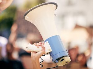 Close-up of a hand holding a megaphone with an out-of-focus crowd in the background.