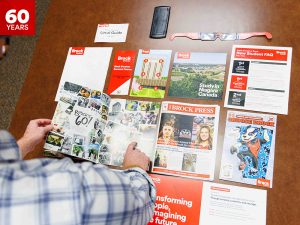 Hands point to a variety of items on a desk, including Brock University magazines and other paper documents.